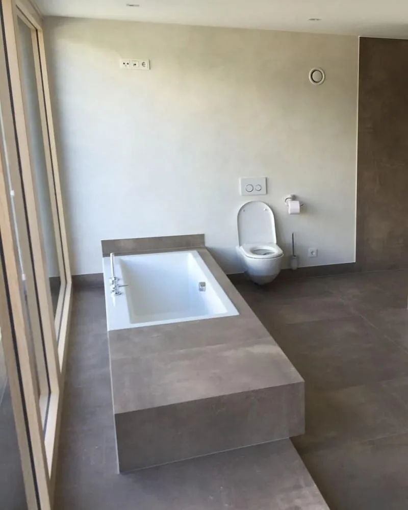 Modernes Badezimmer mit minimalistischem Design. In der Mitte des Raumes befindet sich eine rechteckige Badewanne mit einer hellen, grauen Verkleidung. Im Hintergrund ist ein wandmontiertes WC zu sehen.