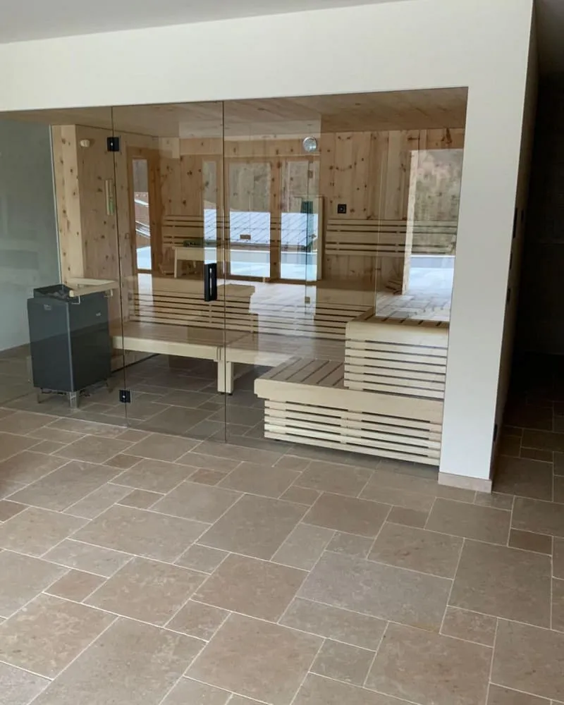 Moderne Sauna mit Glasfront und Holzausstattung. Die Innenwände und Bänke der Sauna bestehen aus hellem Holz, und durch die Glasfront fällt viel Tageslicht in den Raum. Rechts befinden sich gestaffelte Sitzbänke, und links ist ein Saunaofen zu sehen. Der Boden im Außenbereich besteht aus beigen Fliesen, die dem Raum ein elegantes und warmes Ambiente verleihen.